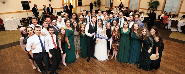 Bride and groom hold a blue Brandeis pennant with a large crowd of guests arrayed around them